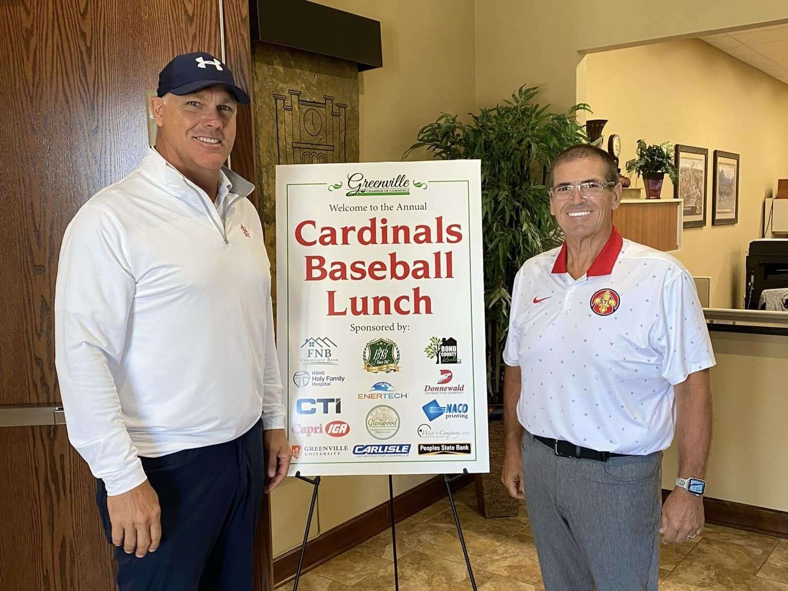 two men in polo shirts and stand smiling indoors by an event welcome sign that includes many sponsoring businesses.